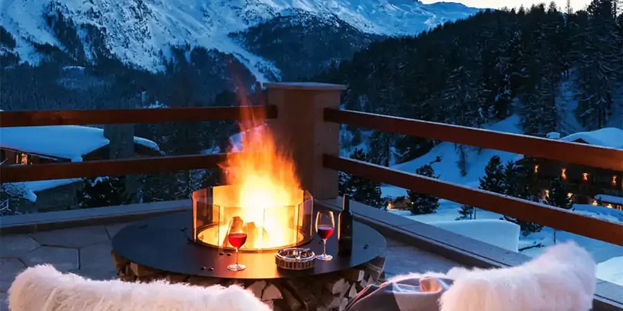 Cozy outdoor seating area with a modern firepit, red wine glasses, and snow-capped mountains in the background during winter.